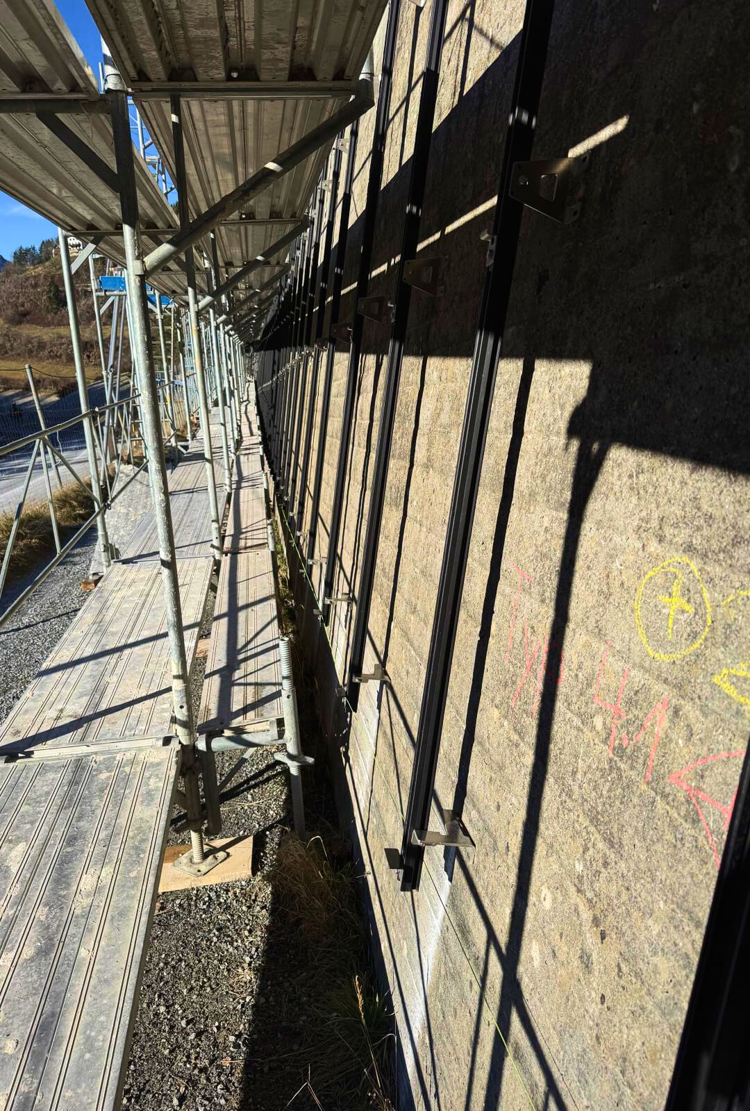 Blick hinter die PV-Module: Montagehalterungen und Tragschienen an der Betonwand, aufgenommen während der Installationsarbeiten vom Gerüst aus. 